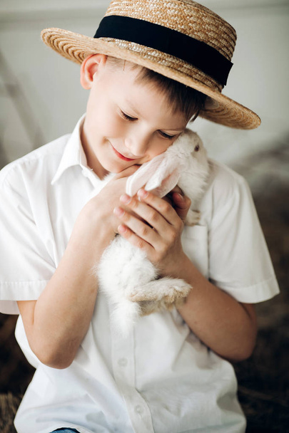 Niño en sombrero de heno manteniendo lindo conejito blanco y sonriendo
 - Foto, imagen