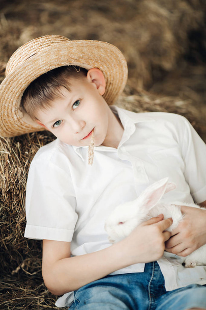 Boy in hay hat keeping cute white bunny and smiling - 写真・画像