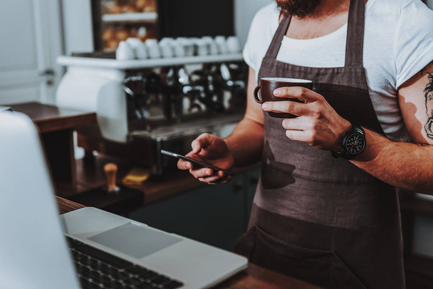 Barista standing with coffee and using modern smartphone - Photo, Image