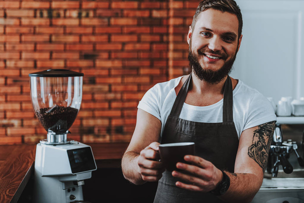 Cheerful barista standing and smiling while holding cup of coffee - Фото, зображення