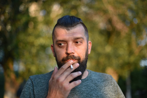 Un hombre con barba fuma un cigarrillo con una camiseta verde en la naturaleza
 - Foto, Imagen