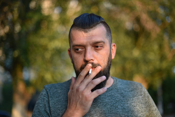 Sakallı bir adam doğada yeşil tişörtle sigara içiyor. - Fotoğraf, Görsel