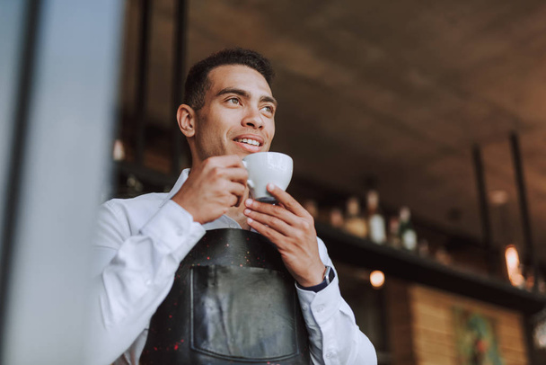 Handsome young man in black apron drinking coffee in cafe - Photo, Image
