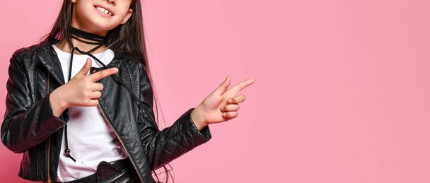 Cropped photo of hipster punk girl - dressed in a leather jacket and skirt, black knee-highs and a white T-shirt, with a funny hairstyle and a makeup star on the face, points to the side - 写真・画像