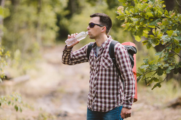 Hiker Drinking Water In Forest. Tired man drinking water from bottle in woodland. Handsome Traveler With Backpack And Flask In Forest. vacation, outdoor lifestyle freedom concept. Adventures hiking - Foto, Imagen