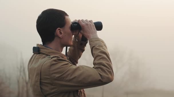 Close up of Young redhead man in khaki coat holding a binoculars looking sunrise and sunset on background. Fog around him. - Footage, Video