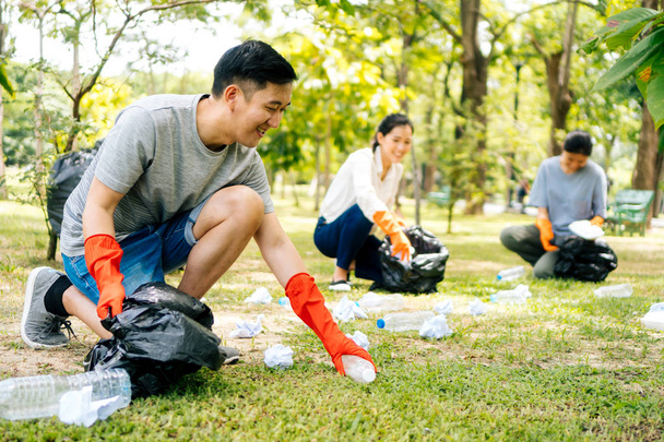 Young Asian man and women wearing orange gloves and collecting trash in garbage bag in the park - Фото, зображення