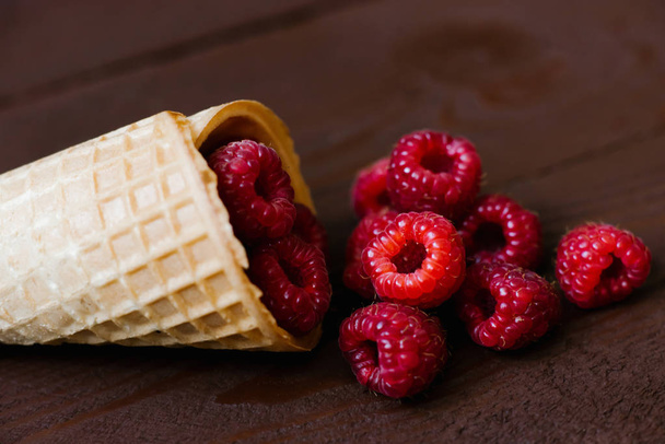 A cascade of fresh raspberries in a waffle cone of ice cream on a brown background. Cafe or restaurant menu. Close up - Photo, Image