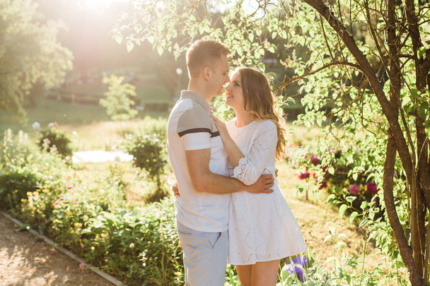 Young pretty couple in love standing in park. Handsome cheerful blonde girl in white dress hugging her boyfriend. Man and woman having fun outdoors - Photo, image