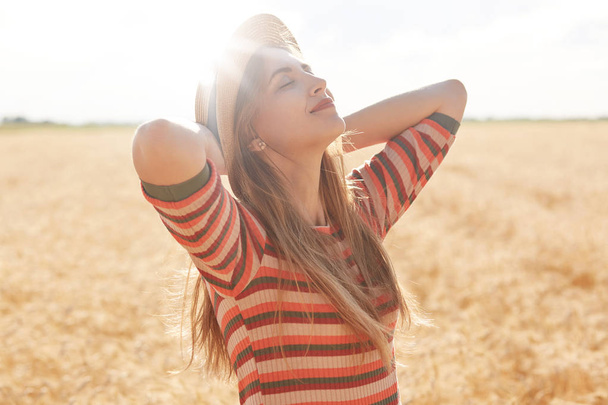 Kültéri shot a boldog fiatal nő csíkos ruhában, és a Sun kalap élvező nap gabonapelyhek területén, női jelentő kezével fel, keres álmodosan fel égen. Természet, nyár, nyaralás és emberikoncepció. - Fotó, kép