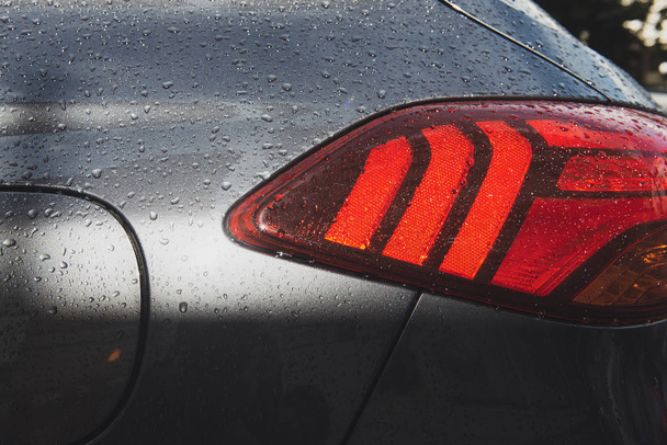 Η πλευρά ενός γκρι αυτοκινήτου. Κοντινά πλάνα σε ένα σύγχρονο οπίσθιο φωτισμό μετά τη βροχή με σταγόνες βροχής. Κόκκινοι ανακλαστήρες που αντανακλούν το φως σε μια ηλιόλουστη μέρα. - Φωτογραφία, εικόνα