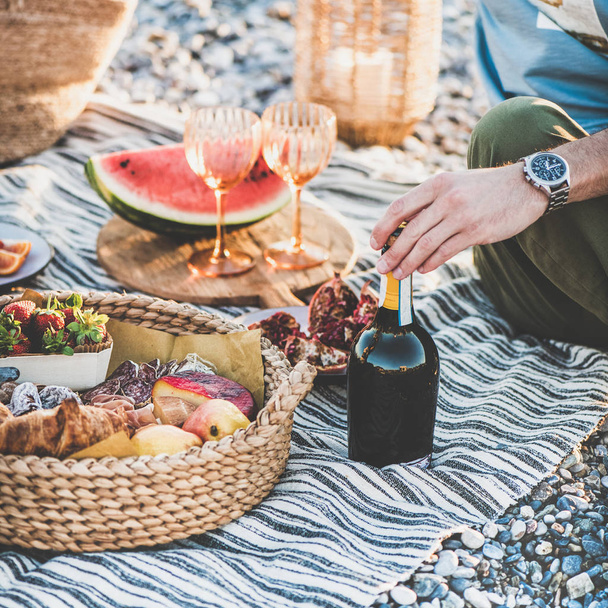Picnic de verano al atardecer. Pareja joven sentada en manta teniendo picnic fin de semana al aire libre en la playa con fruta fresca de temporada, bandeja de sabrosos aperitivos y botella de vino espumoso, cosecha cuadrada
 - Foto, imagen