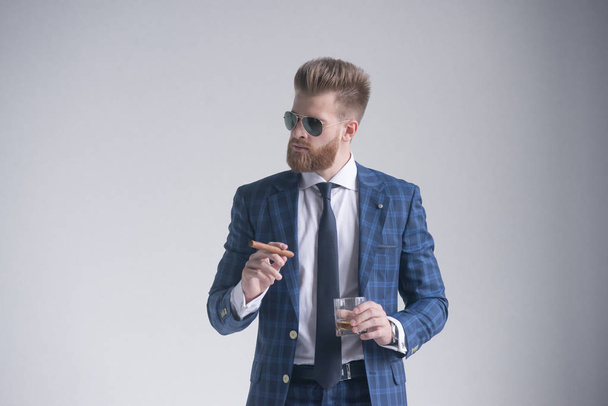 Hombre guapo y confiado sostiene un cigarro y un vaso de whisky y mira hacia otro lado mientras está de pie en el interior
 - Foto, Imagen