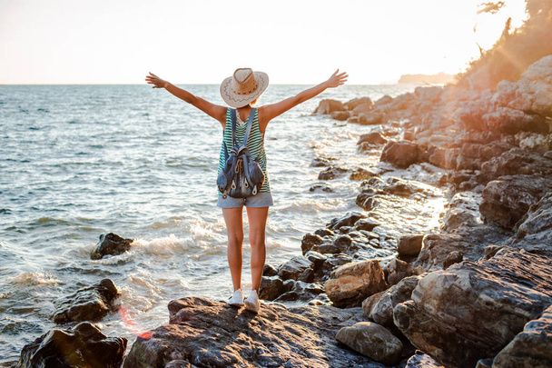 A young hipster woman in a hat and a rukzak with her hands up, standing on top of a cliff and looking at the sea at sunset. - Fotografie, Obrázek
