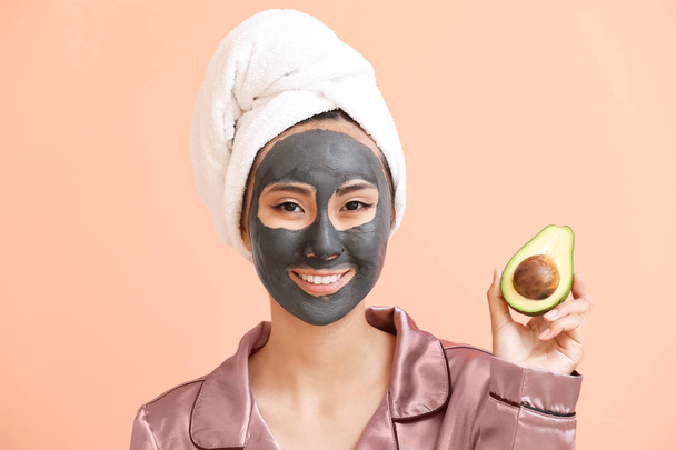 Beautiful Asian woman with facial mask and avocado on color background - Фото, изображение