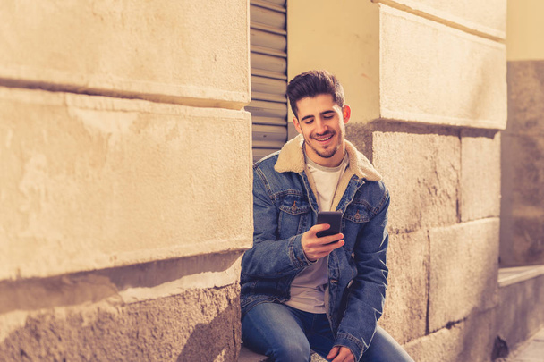 Young attractive college man in his twenties happy talking and chatting with friend on smart mobile phone on campus or city stairs. In communication, technology internet and travel around europe. - Fotó, kép