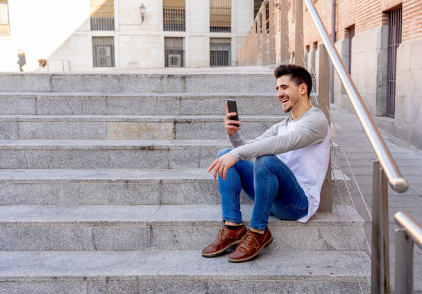 Jonge aantrekkelijke college man in zijn twintiger jaren gelukkig praten en chatten met een vriend op slimme mobiele telefoon op campus of stad trappen. In communicatie, technologie internet en reizen rond Europa. - Foto, afbeelding