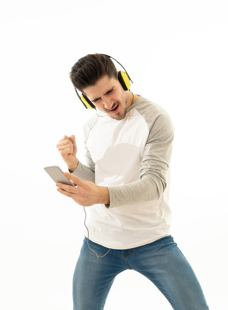 Portrait of happy young man listening to music in headphones changing songs and tracks on smartphone and singing and dancing. Isolated white background. In positive emotions, youth happiness concept. - Фото, зображення