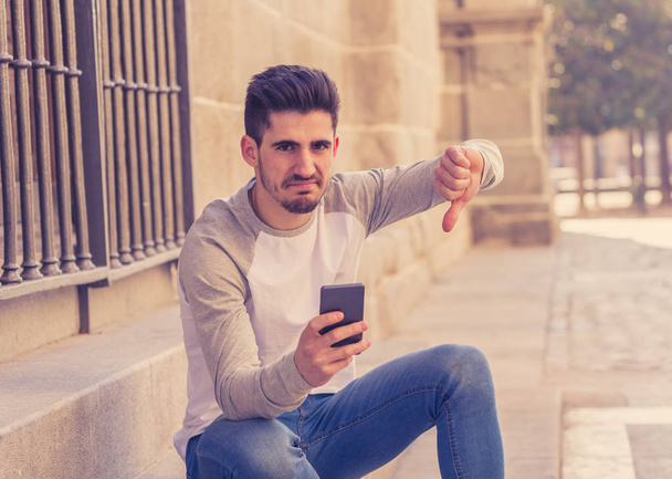 Portrait of young student man reacting negative to post in social media network or dating app with dislike showing thumb down. In social network rating likes, reactions and reassurance youth problems. - Foto, imagen