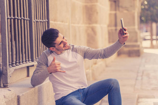Excited trendy young man taking a selfie on smart mobile phone on holidays in european city street or in university campus outside. In holidays and technology, connection and social media concept. - Foto, imagen