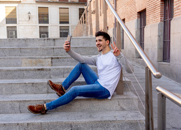 Excited trendy young man taking a selfie on smart mobile phone on holidays in european city street or in university campus outside. In holidays and technology, connection and social media concept. - Fotografie, Obrázek