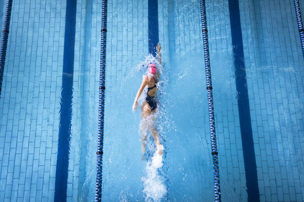 Korkea kulma näkymä valkoihoinen nainen yllään uimapuku ja vaaleanpunainen uimalakki tekee freestyle aivohalvaus uima-altaassa
 - Valokuva, kuva