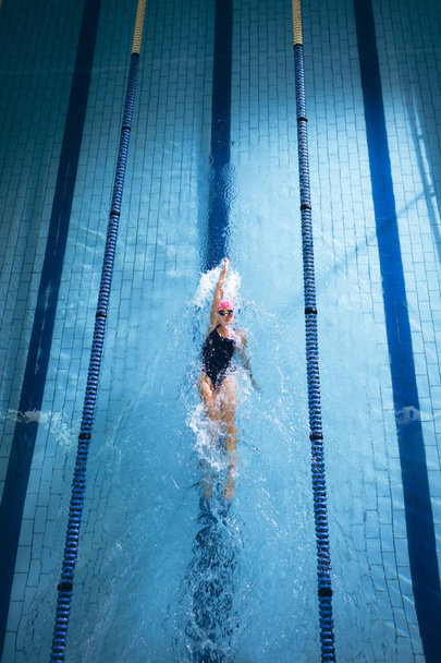 Vista de alto ângulo de uma mulher caucasiana vestindo um boné de natação rosa e óculos fazendo um golpe nas costas em uma piscina
 - Foto, Imagem