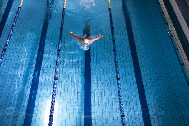 Widok z dużym kątem mężczyzn kaukaski pływak ubrany w białą czapkę pływania robi skok motyla w basenie - Zdjęcie, obraz