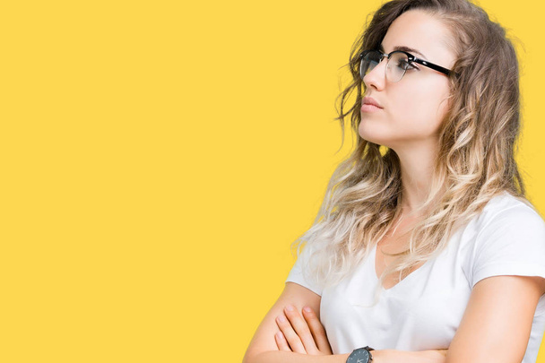 Beautiful young blonde woman wearing glasses over isolated background smiling looking to the side with arms crossed convinced and confident - Photo, Image