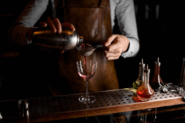 Male bartender pouring a red alcoholic drink from the steel shaker through the sieve - Foto, Imagen