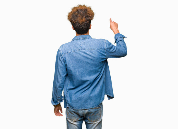 junger gutaussehender Mann mit Afro-Haaren in Jeansjacke posiert rückwärts und zeigt mit der Fingerhand nach hinten - Foto, Bild