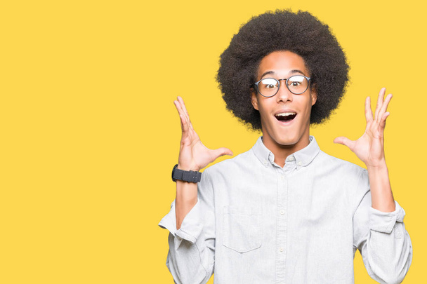 Молодой африканский американец с афроволосами в очках празднует сумасшедший и поражен успехом с поднятыми руками и открытыми глазами, крича взволнованным. Концепция победителя
 - Фото, изображение