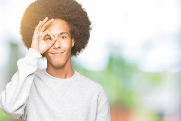 スポーティなスウェットシャツを着たアフロヘアの若いアフリカ系アメリカ人男性は、手を笑顔でOKジェスチャーを行い、幸せな顔で指を通して目を見る. - 写真・画像