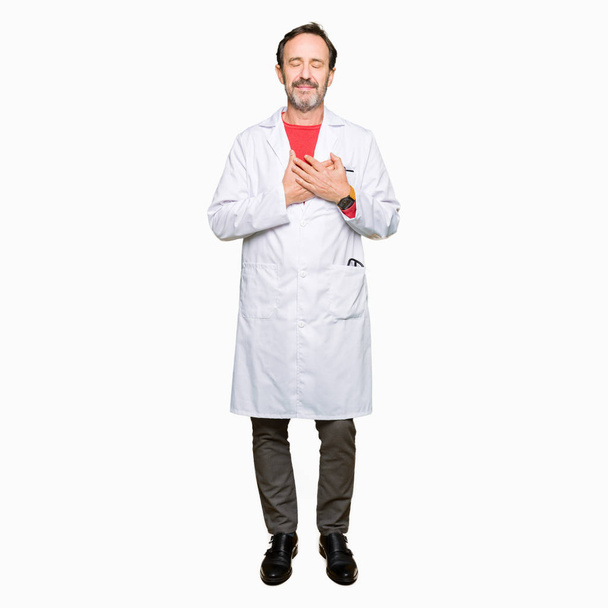 Middle age therapist wearing white coat smiling with hands on chest with closed eyes and grateful gesture on face. Health concept. - Photo, Image