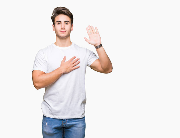 Молодой красивый мужчина в белой футболке на изолированном фоне, ругаясь рукой на груди и открытой ладони, дает клятву верности
 - Фото, изображение