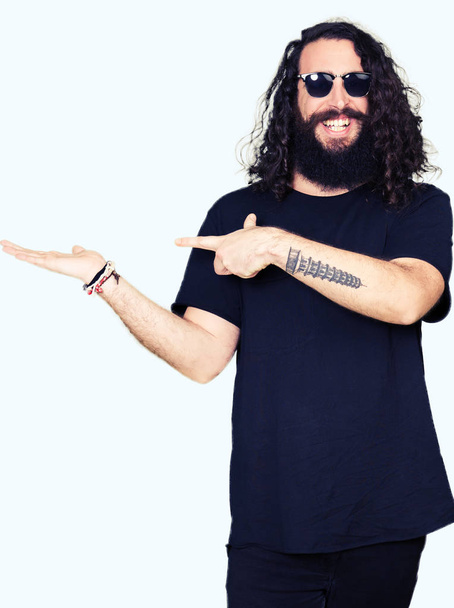 Jonge hipster man met lange haren en baard dragen van een zonnebril verbaasd en lachend naar de camera tijdens het presenteren met hand en met de vinger te wijzen. - Foto, afbeelding