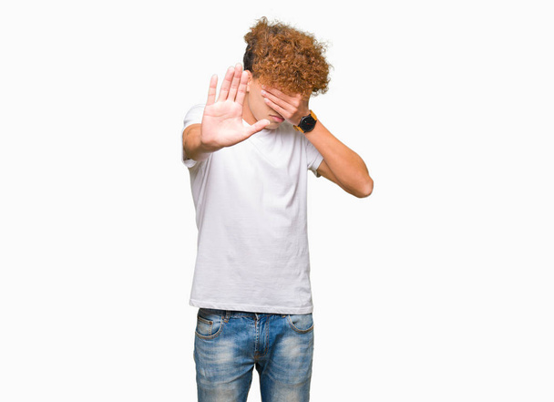 Young handsome man with afro hair wearing casual white t-shirt covering eyes with hands and doing stop gesture with sad and fear expression. Embarrassed and negative concept. - 写真・画像