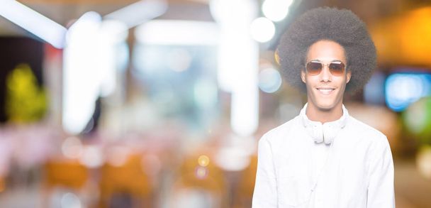 junger afrikanisch-amerikanischer Mann mit Afro-Haaren, Sonnenbrille und Kopfhörer mit einem fröhlichen und kühlen Lächeln im Gesicht. Glück gehabt. - Foto, Bild