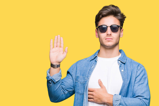 Mladý pohledný muž nosí sluneční brýle přes samostatný pozadí nadávky s rukou na hrudi a otevřené dlaně, aby slib slib věrnosti - Fotografie, Obrázek