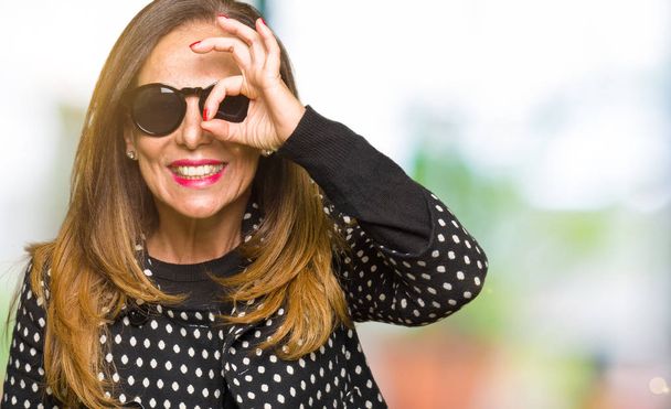 Красива жінка середнього віку в сонцезахисних окулярах робить нормальний жест з посмішкою руки, очі дивляться через пальці з щасливим обличчям
. - Фото, зображення