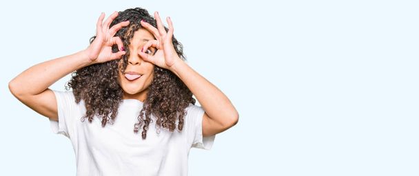 Young beautiful woman with curly hair wearing white t-shirt doing ok gesture like binoculars sticking tongue out, eyes looking through fingers. Crazy expression. - Photo, image