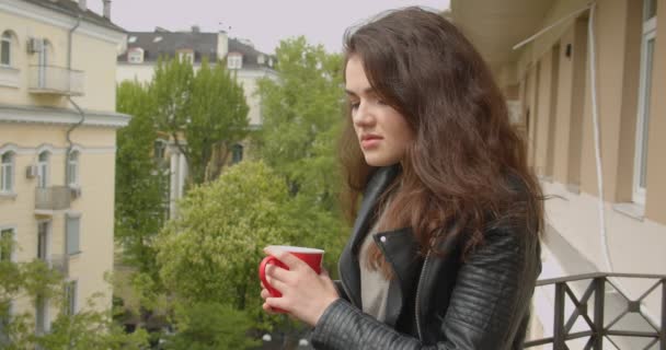 Young brunette girl in leather jacket standing on balcony enjoying the spectacular green city view and drinking hot tea. - Πλάνα, βίντεο