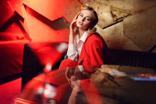 Photo of young blonde woman in red jacket sitting on sofa at table - Foto, imagen