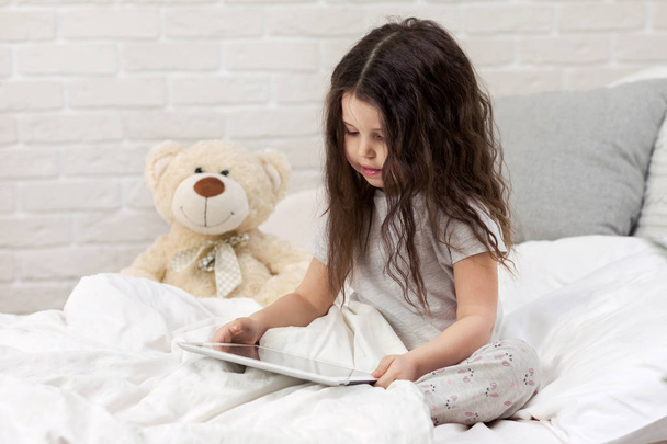 cute little girl lies in bed uses digital tablet. - Фото, изображение