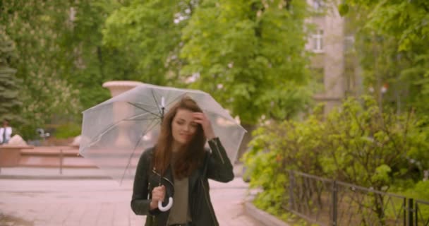 Portrait of cute of caucasian brunette female walking with the umbrella smiles into camera in the green park. - Filmati, video