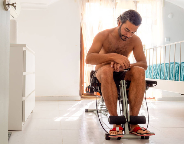 Brunet man on household gym chair during workout in bedroom - Photo, Image