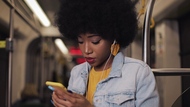 Portrait of serious african american women in headphones listening to music and browsing on mobile phone in public transport. City lights background. Slow motion. - Felvétel, videó