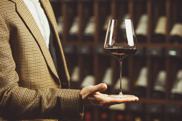 Glass of red wine in the sommelier hand on the cellar with bottles backgrounds. - Fotó, kép