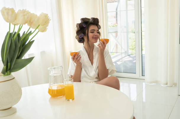 beautiful young brunette woman in hair curlers holding oranges in both hands in bright kitchen - Fotó, kép