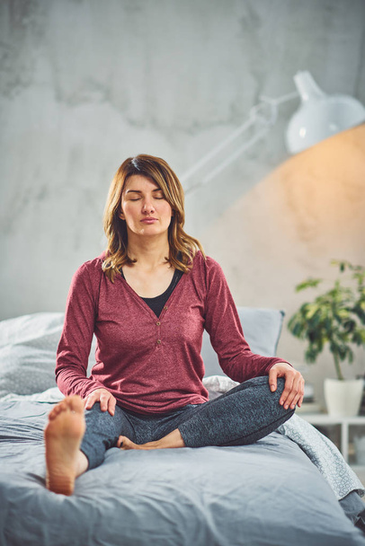 Affascinante bruna caucasica seduta sul letto in camera da letto e facendo esercizi di yoga
. - Foto, immagini